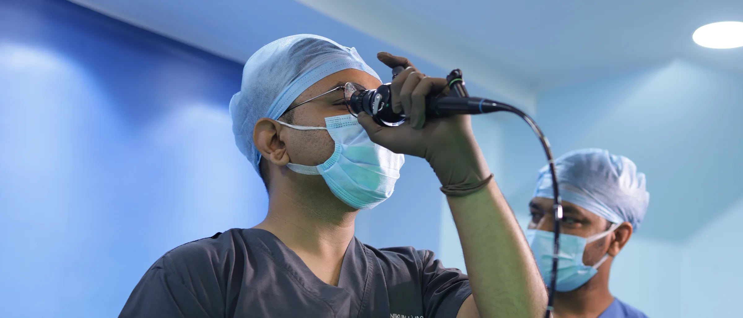 A surgeon holding a flexible endoscope up to his eye, a colleague watching beside him.