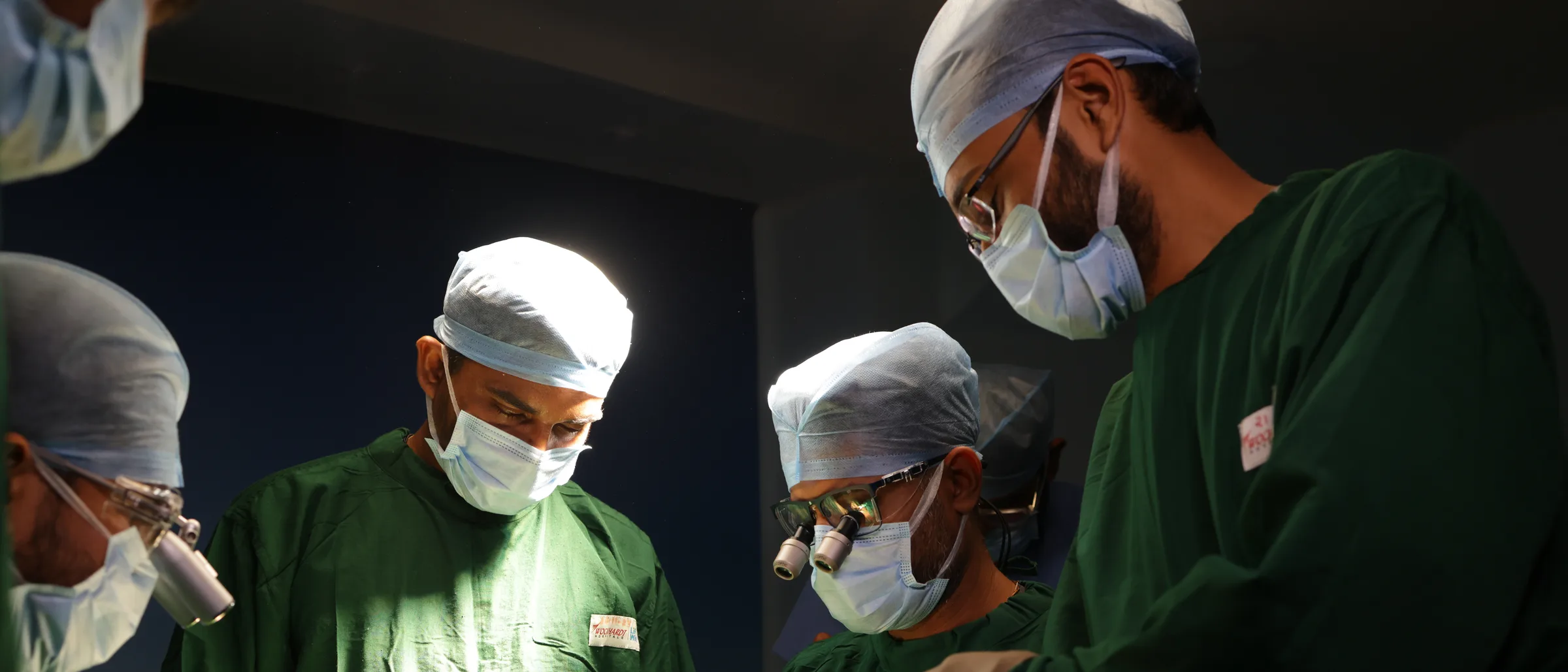 Four surgeons in green gowns bowed over the operating table, seen from below against a bare wall.