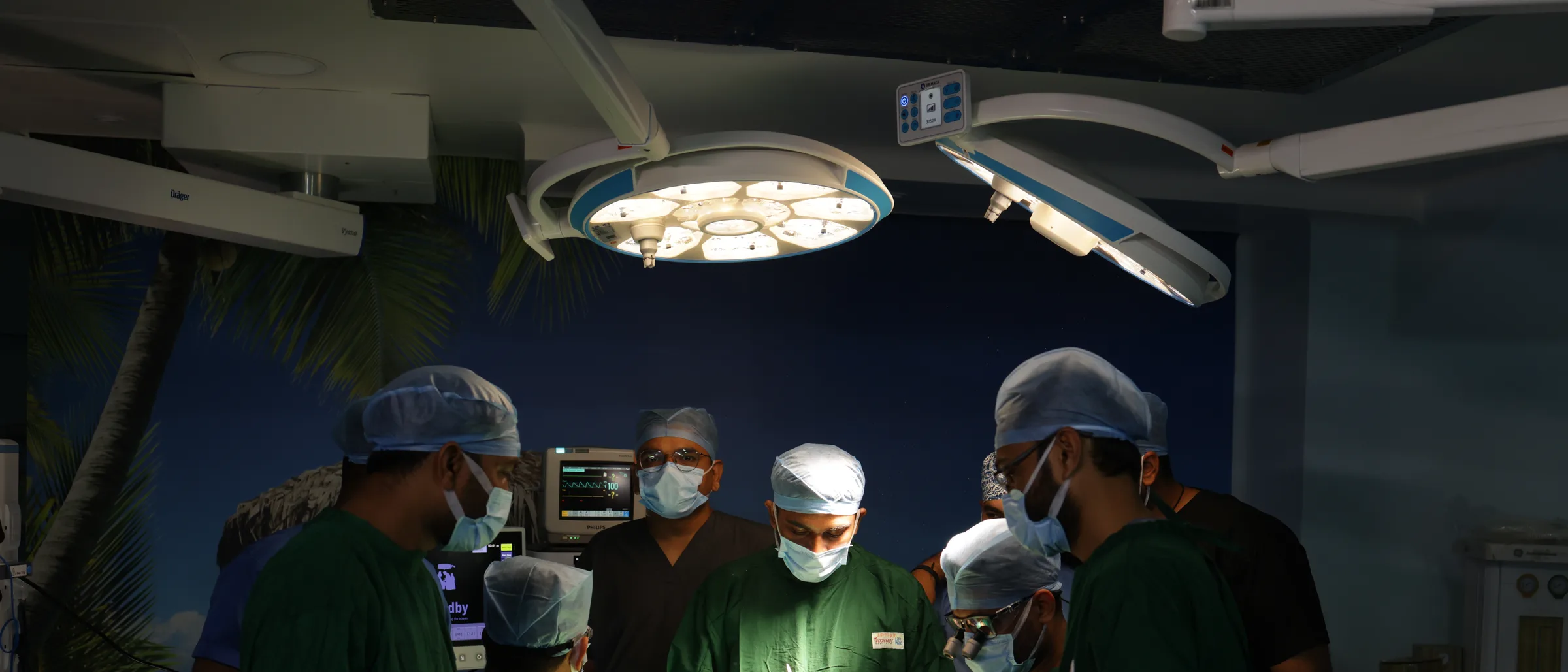 The operating theatre from the foot of the table: two overhead lamps and eight of the team round one draped patient.