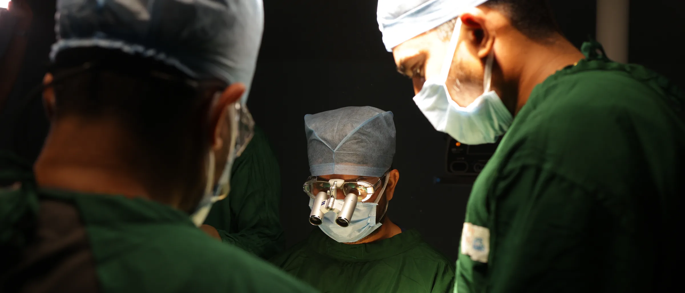 Three surgeons leaning in close over the operating table, two of them seen from behind.