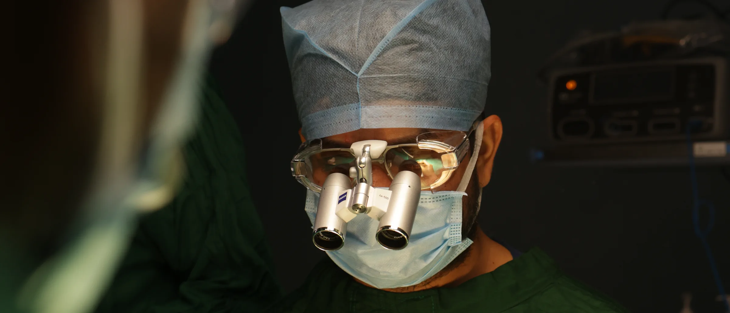 A surgeon in surgical loupes, capped and masked, looking down at his work in the operating theatre.