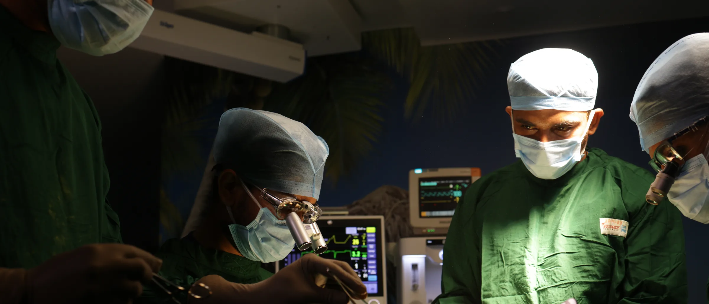 A surgeon wearing magnifying loupes working with fine forceps, a monitor showing a live trace behind him and a gowned colleague opposite.