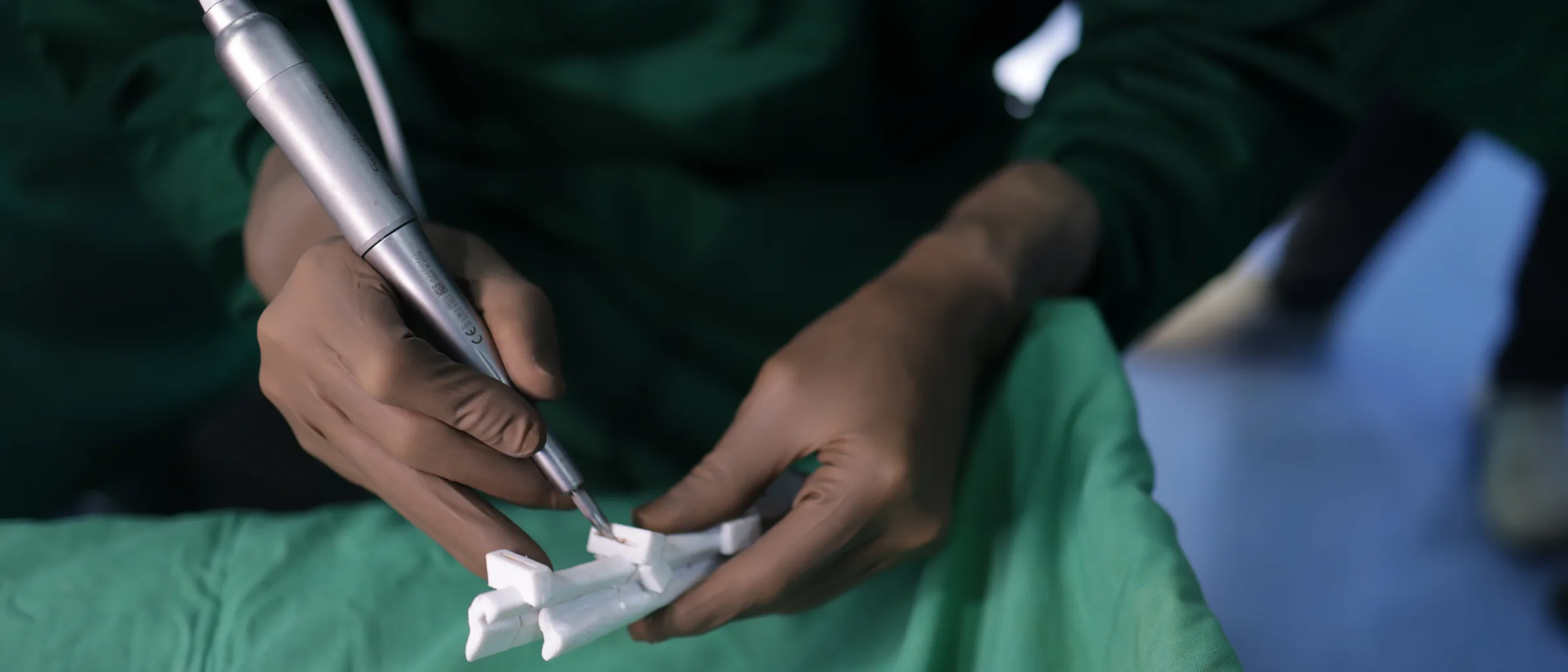 A surgeon's gloved hands over green drapes, using a powered saw to cut a white model into angled segments.