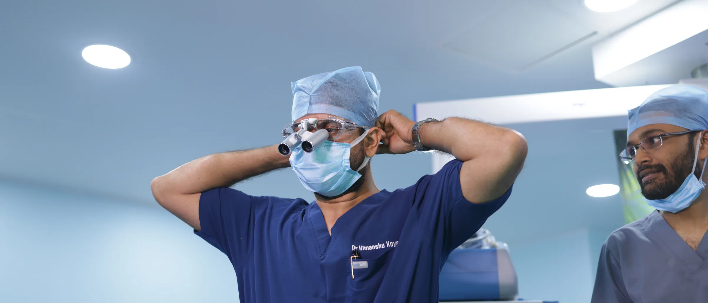 A surgeon in navy scrubs fitting magnifying loupes over his mask before an operation, a colleague standing to his right.