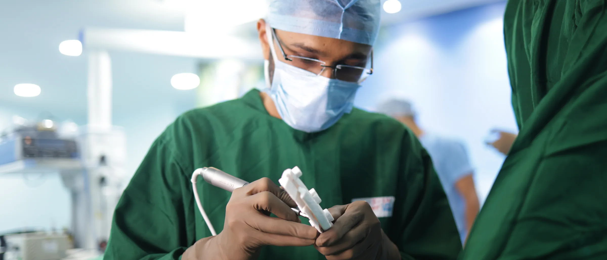 A surgeon's gloved hands checking an instrument before a procedure, his face masked above them.