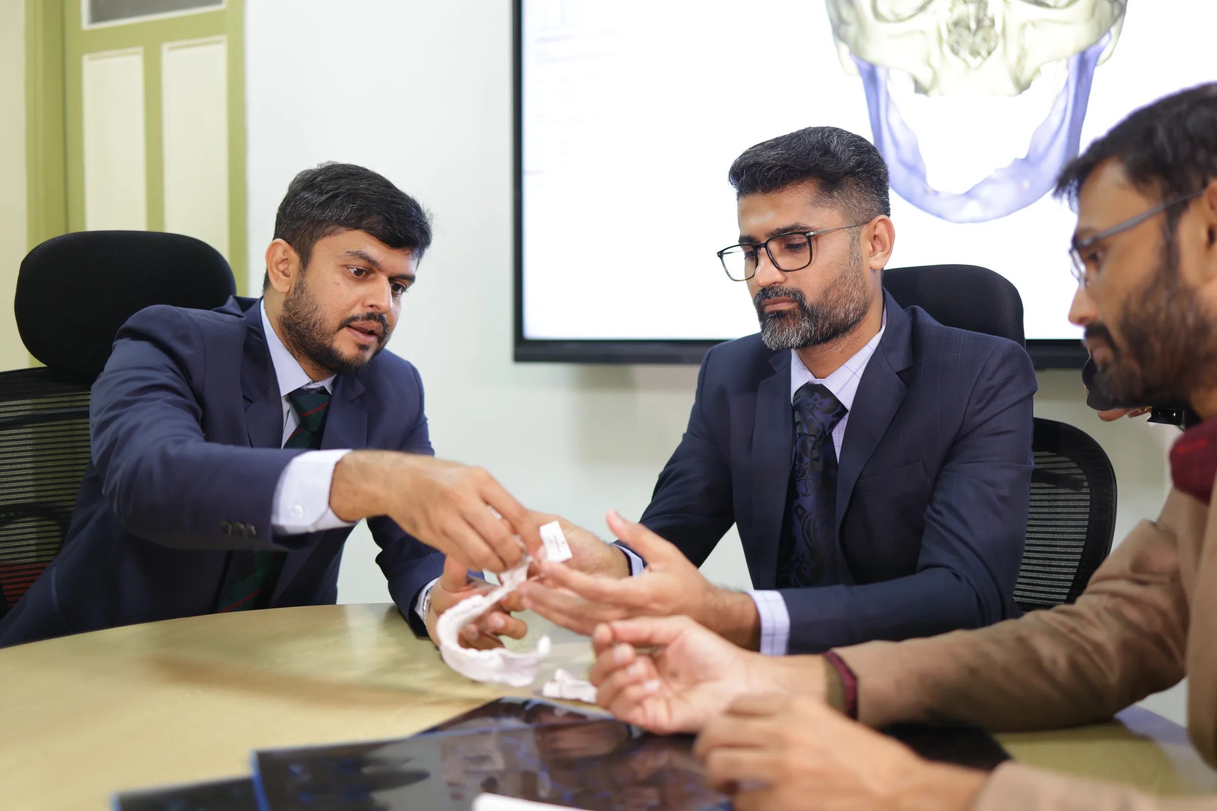 Two cancer surgeons at the tumour board passing a 3D-printed model of a jaw between them, with the scan it was made from on the screen behind.