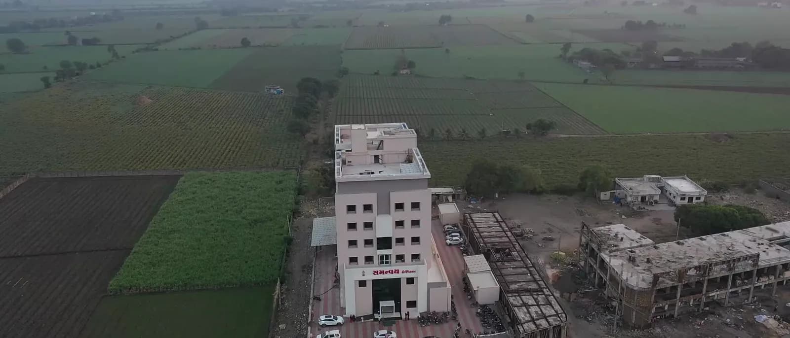Samanvay Hospital from the air at dusk, cars and motorcycles parked along the road at its entrance and green fields on every side.