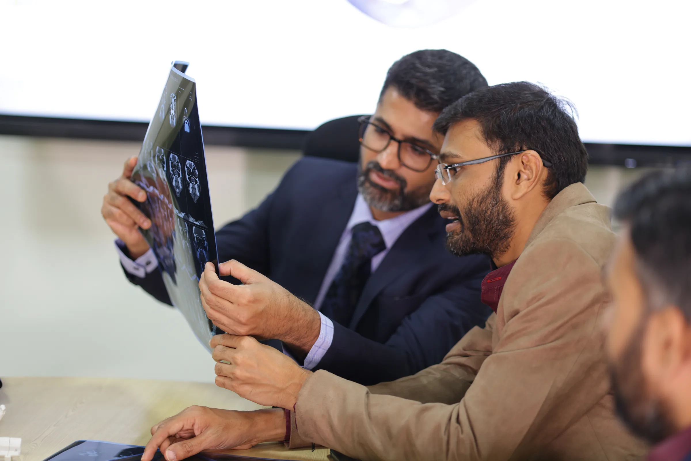 A cancer surgeon holding a scan film up to the light while a colleague reads it beside him.